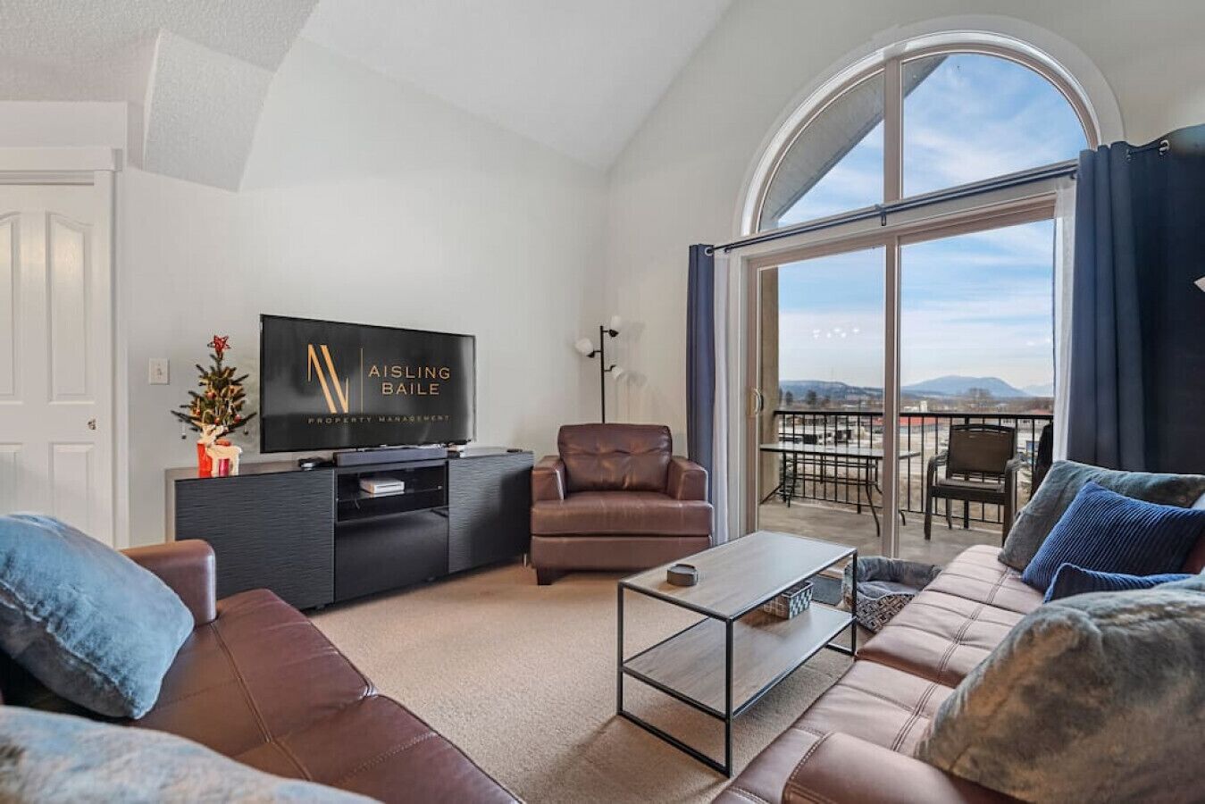 A modern living room with a large TV, leather sofas, a coffee table, and a balcony with a view.