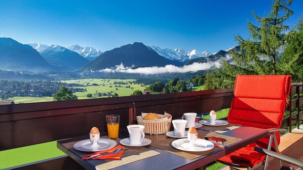 Mesa de café da manhã com pratos, xícaras e uma cesta, com vista para uma paisagem montanhosa.