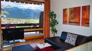Uma sala de estar com uma grande janela com vista para montanhas, uma televisão de tela plana, um sofá e um vaso de planta.