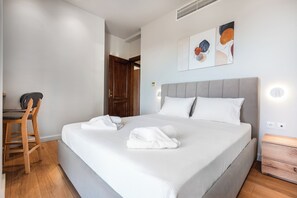 A neatly made bed with white linens, a wooden chair, a bedside table, and a door in the background.