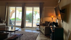 A spacious living room with a wooden ceiling, a sofa, a coffee table, and a TV. Large windows with curtains offer a view of palm trees and a pool. - New Private Pool, Private Beach and Flat, Private House, what else do you need? (Whymms)