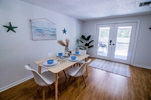 A dining area with a wooden table set for four, white chairs, a plant, and a door leading outside.