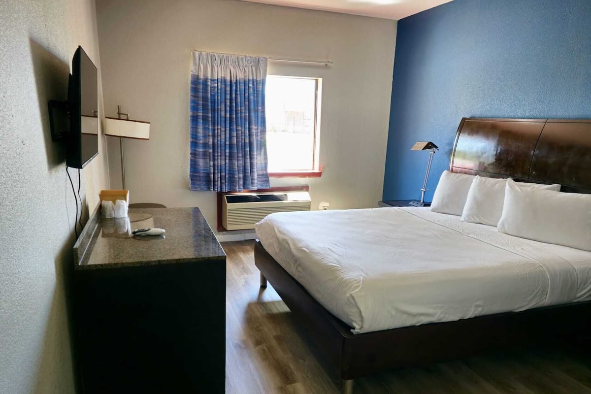 Une chambre d’hôtel comprenant un lit, un bureau, un téléviseur à écran plat, une fenêtre avec des rideaux et une table de chevet avec une lampe.