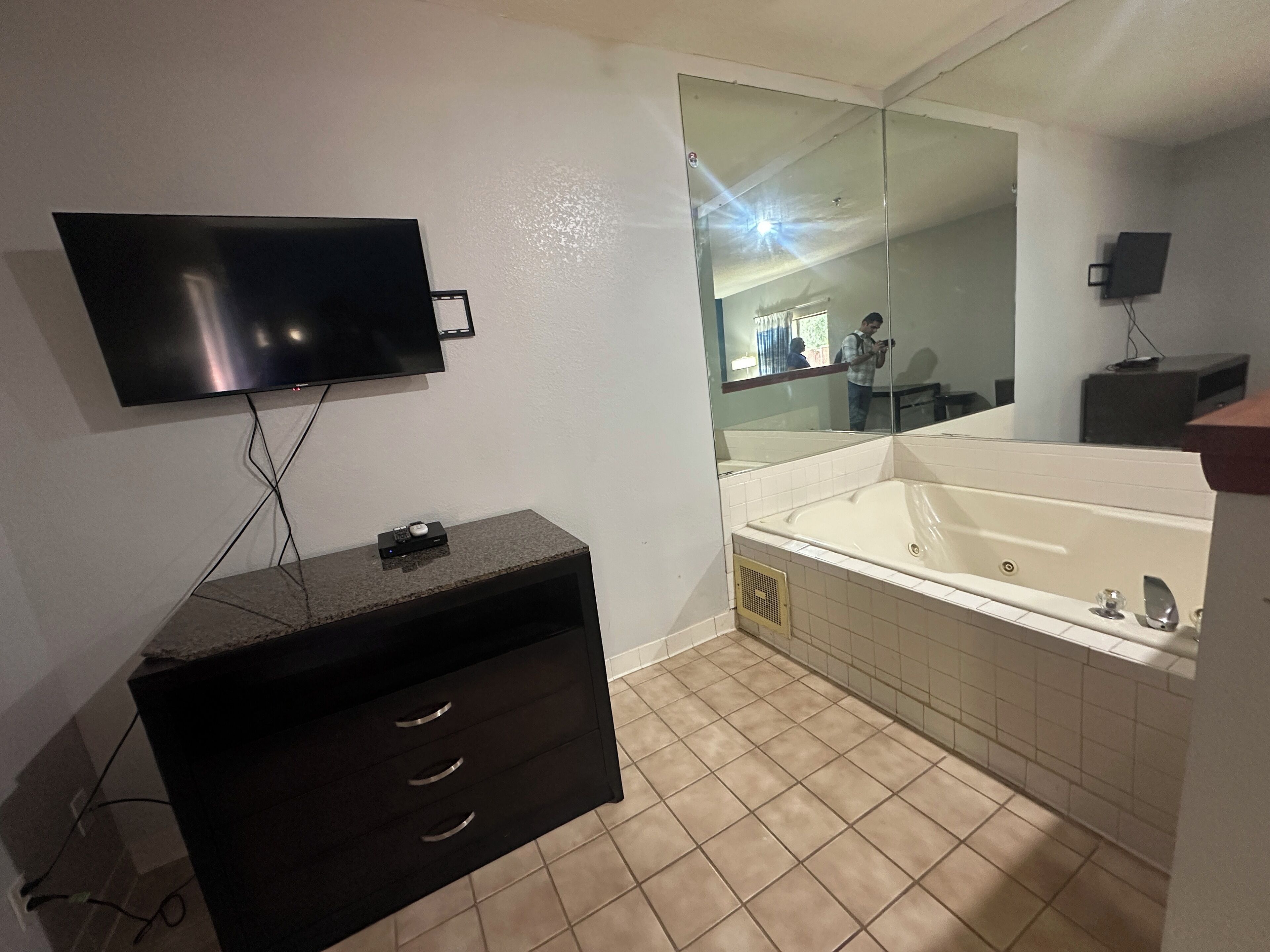 A modern bathroom with a large mirror, a flat-screen TV mounted above a dark wooden vanity, and a freestanding bathtub.
