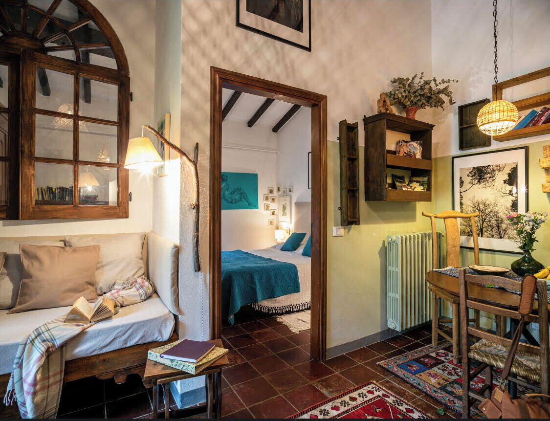A cozy room with a bed, a sofa, a wooden chair, and a table with books. There is a door leading to another room with a bed and a painting on the wall.