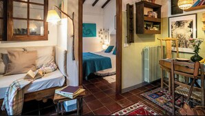 A cozy room with a bed, a sofa, a wooden chair, and a table with books. There is a door leading to another room with a bed and a painting on the wall. - Cozy little retreat in wonderful location, with pool (località Fornello, Pontassieve)