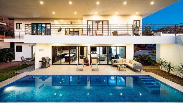 Una moderna casa a due piani con una grande piscina, area salotto esterna e un balcone.
