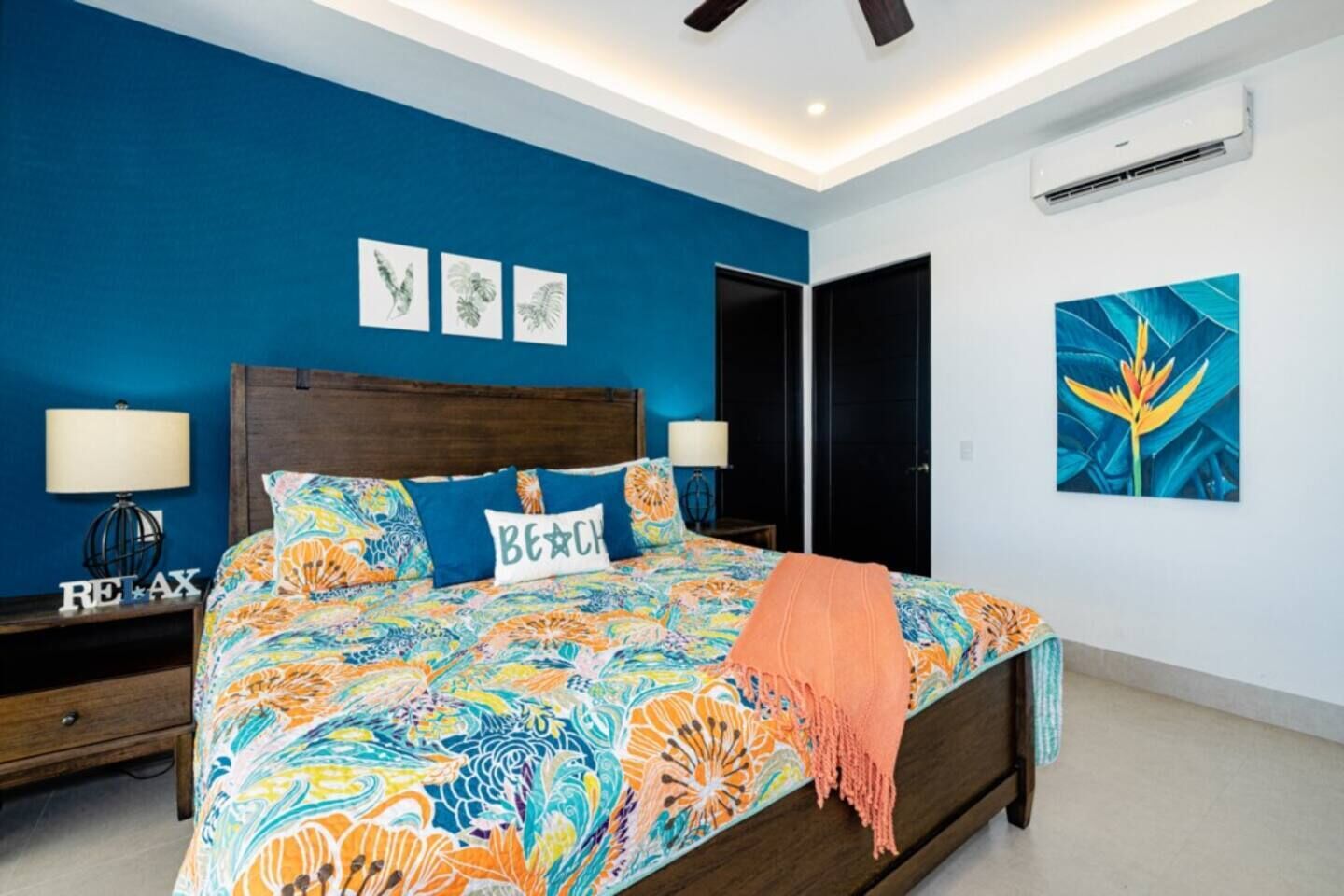 A bedroom with a large bed, a nightstand with a lamp, a ceiling fan, and artwork on the walls.