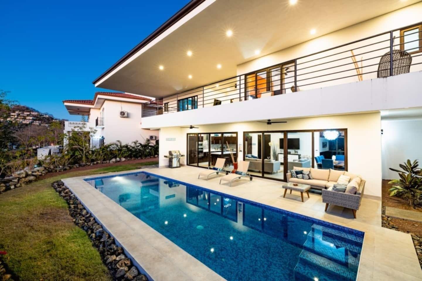 Una casa moderna de dos pisos con piscina, mobiliario exterior y un balcón.