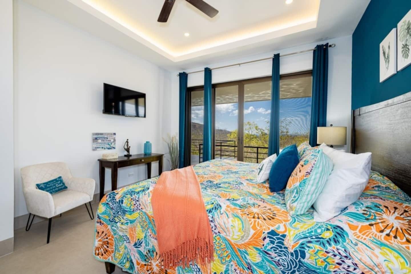 A bedroom with a large bed, a chair, a desk, a TV, and a view of the outdoors.