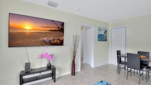 A living room with a large flat-screen TV mounted on the wall, a dining table with chairs, a blue rug, and a vase with dried reeds. - Beautiful spacious 2bd, w/d, bbq, yard, pets allowed, free parking  (Delray Beach)
