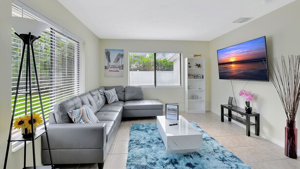 A modern living room with a grey sectional sofa, a white coffee table, a TV on the wall, and a large window with blinds. - Beautiful spacious 2bd, w/d, bbq, yard, pets allowed, free parking  (Delray Beach)