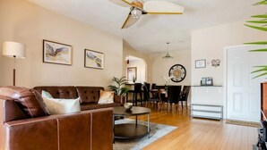 A living room with a brown leather sofa, a coffee table, and a ceiling fan. There are framed pictures on the wall and a dining area in the background. - National Park Home Base Between Miami & The Keys (Homestead)