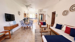 A spacious living area with a sofa, chairs, a coffee table, and a TV. There is a kitchen in the background with a dining table and chairs. - Modern 2BR in Cabarete • Pool • Beach Walk (Cabarete)