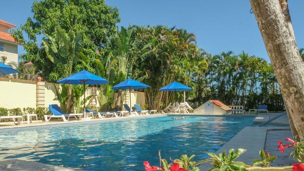 A swimming pool with lounge chairs and blue umbrellas, surrounded by palm trees and a clear sky. - Modern 2BR in Cabarete • Pool • Beach Walk (Cabarete)