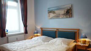 A neatly made bed with a headboard, two bedside lamps, a window with curtains, and a painting of a beach scene on the wall. - Apartment Famli Relax **** Villa Störtebeker in Göhren (Göhren)