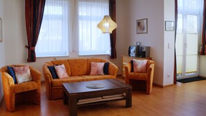 A living room with a sofa set, a coffee table, and a lamp. There are windows with curtains, a television, and a door with a mat. - Apartment Famli Relax **** Villa Störtebeker in Göhren (Göhren)