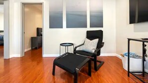 Un salon moderne avec un plancher en bois, un fauteuil noir agrémenté d’un coussin gris, une petite table d’appoint noire et un grand téléviseur à écran plat fix é au mur.