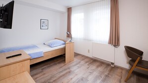 Une petite chambre à coucher, bien rangée, avec un lit simple, un bureau, une chaise, un téléviseur fixé au mur et une fenêtre avec des rideaux.