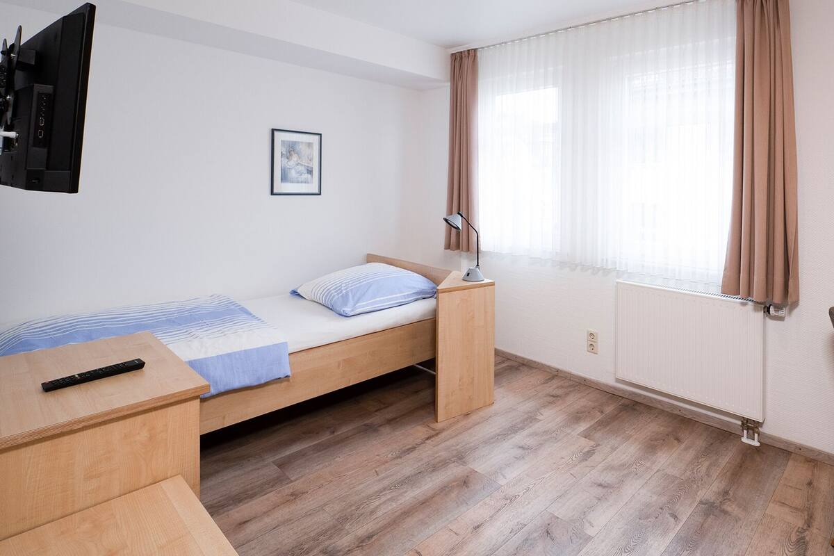 Un dormitorio pequeño y ordenado con una cama individual, un escritorio, una silla, un televisor montado en la pared y una ventana con cortinas.