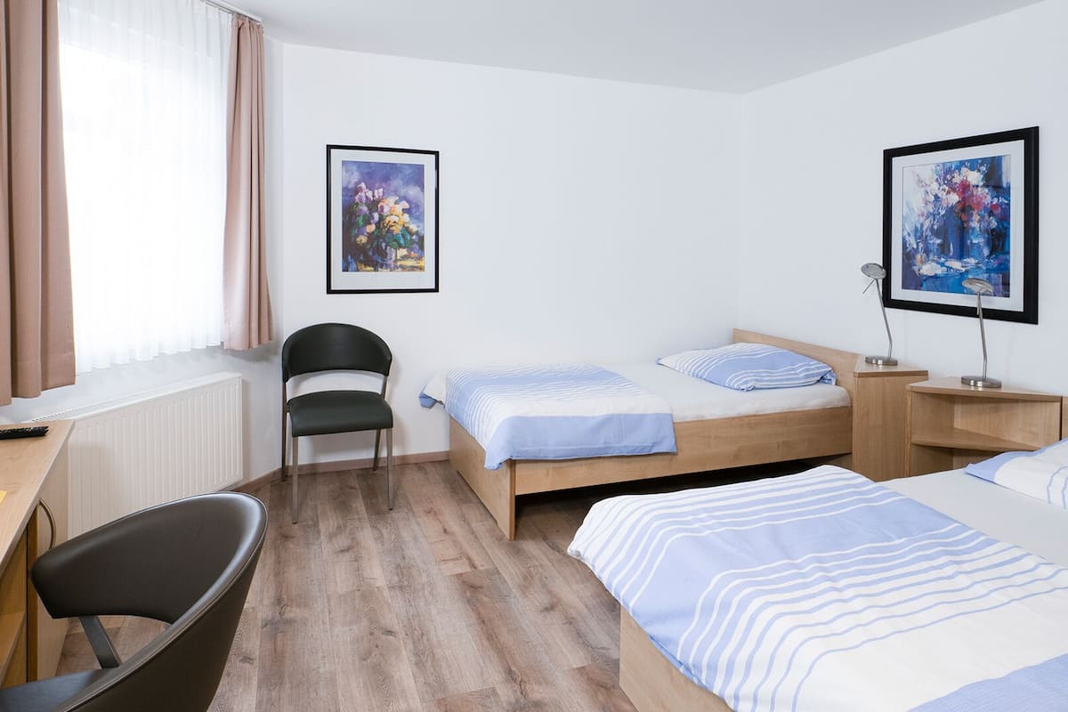 Habitación de hotel con cama, escritorio, silla, ventana con cortinas y dos cuadros en la pared.
