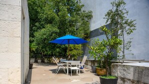 Um pátio externo com mesa e cadeiras, um guarda-sol azul e vasos de plantas.