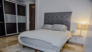 Cama bem arrumada com cabeceira cinzenta, luminária de cabeceira e guarda-roupa espaçoso com portas deslizantes.