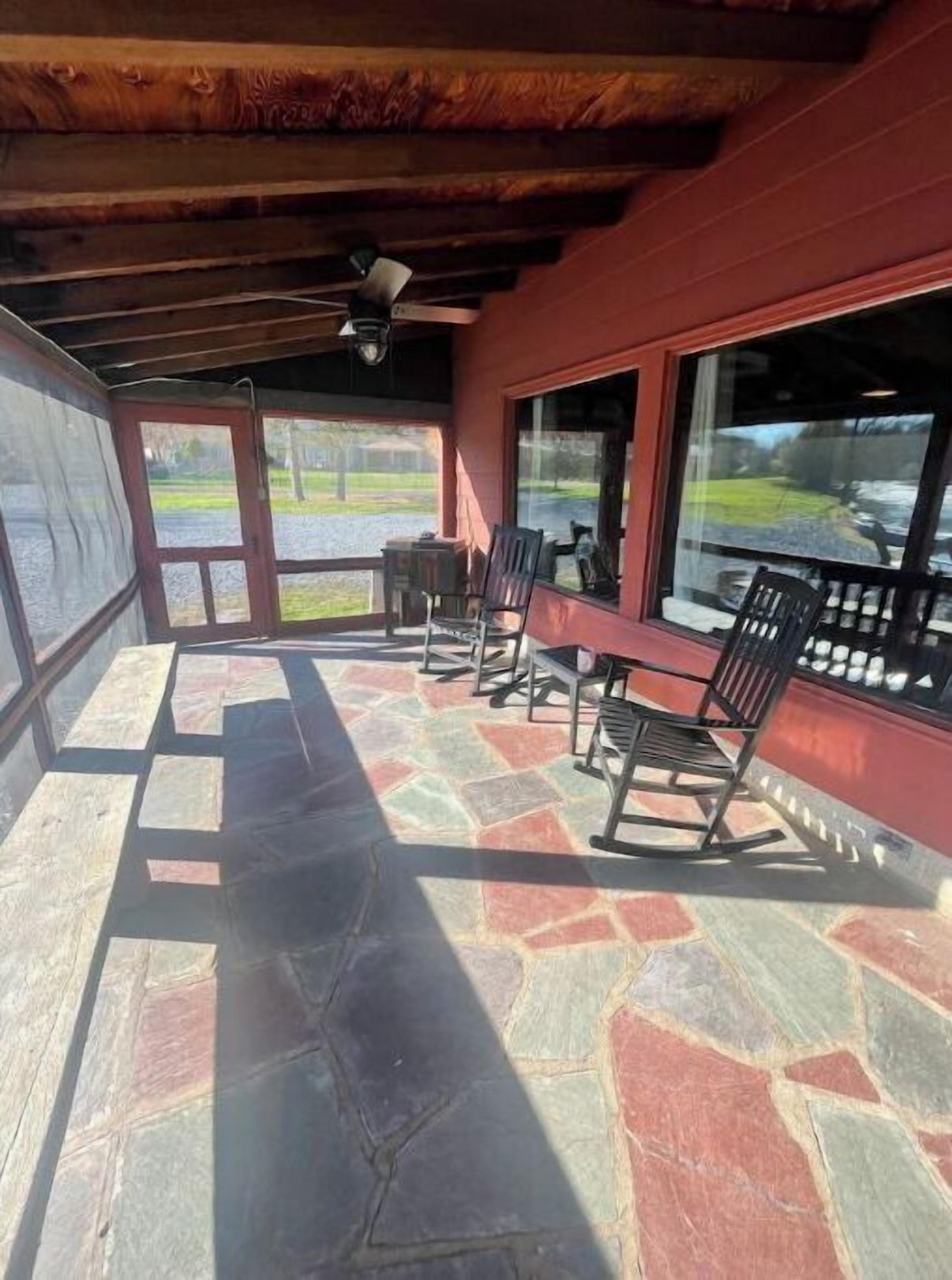 A covered patio with stone flooring, wooden chairs, and large windows offering views of a green landscape.