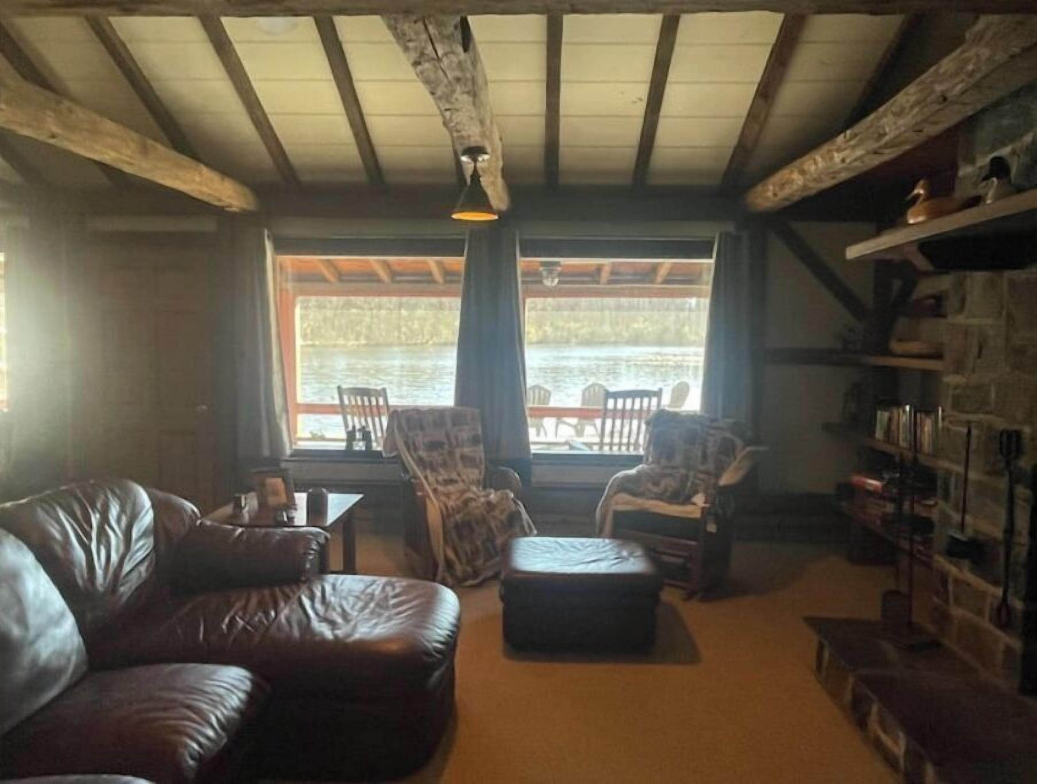 Ein gemütliches Wohnzimmer mit einer Ledersofa, einem kleinen Hocker und einem Esstisch mit Stühlen. Eine Bücherwand ist rechts sichtbar, und das Zimmer hat eine rustikale Holzbalkendecke.