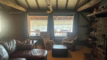 Ein gemütliches Wohnzimmer mit einer Ledersofa, einem kleinen Hocker und einem Esstisch mit Stühlen. Eine Bücherwand ist rechts sichtbar, und das Zimmer hat eine rustikale Holzbalkendecke.