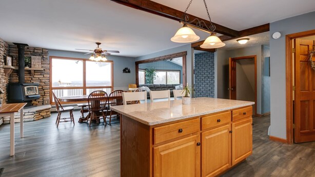 A spacious kitchen with a large island, wooden cabinets, and a stone fireplace. There is a dining area with a table and chairs, and a view of the outdoors through large windows. - Kettle Creek Hills Lodge: Exceptional Views! (Elk Township)