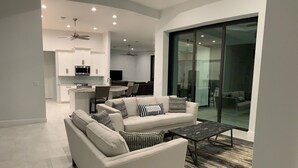 A modern living room with a sofa, a coffee table, and a patterned rug. There is a kitchen in the background with white cabinets and a dining area with a couch and chairs. - Entire 4 Bedroom W/Pool Access (Marco Island)