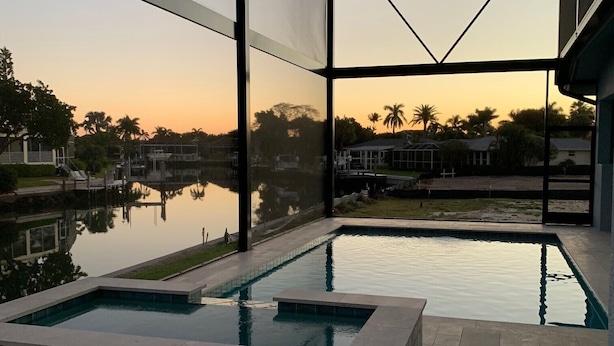 A modern outdoor pool with a geometric glass enclosure, surrounded by a calm water body and palm trees. - Entire 4 Bedroom W/Pool Access (Marco Island)