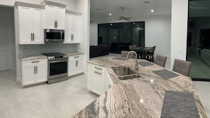 A modern kitchen with white cabinets, a built-in microwave, a stove, and a large granite island with a sink. - Entire 4 Bedroom W/Pool Access (Marco Island)