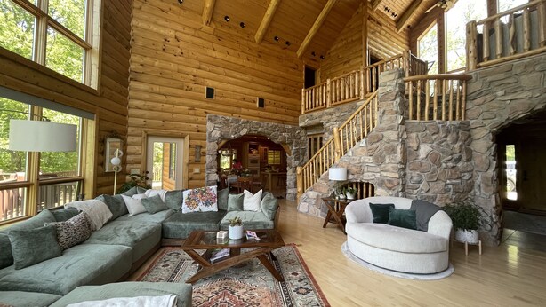 A spacious log cabin interior with a large living area, a stone fireplace, and a wooden staircase.