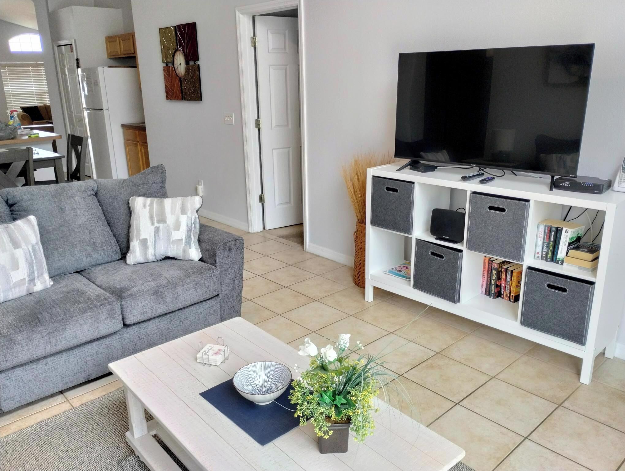 Ein modernes Wohnzimmer mit einem grauen Sofa, einem Couchtisch mit einer Schale und Blumen, einem Fernseher auf einem weißen Schrank und einem Bücherregal mit Aufbewahrungsboxen.