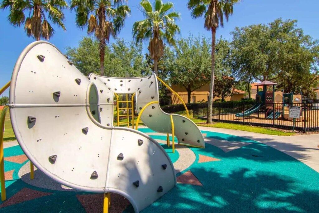 A playground with climbing structures and a slide, surrounded by palm trees and a fenced area.