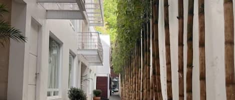 A narrow pathway flanked by buildings with a row of bamboo plants on one side and a metal railing on the other.