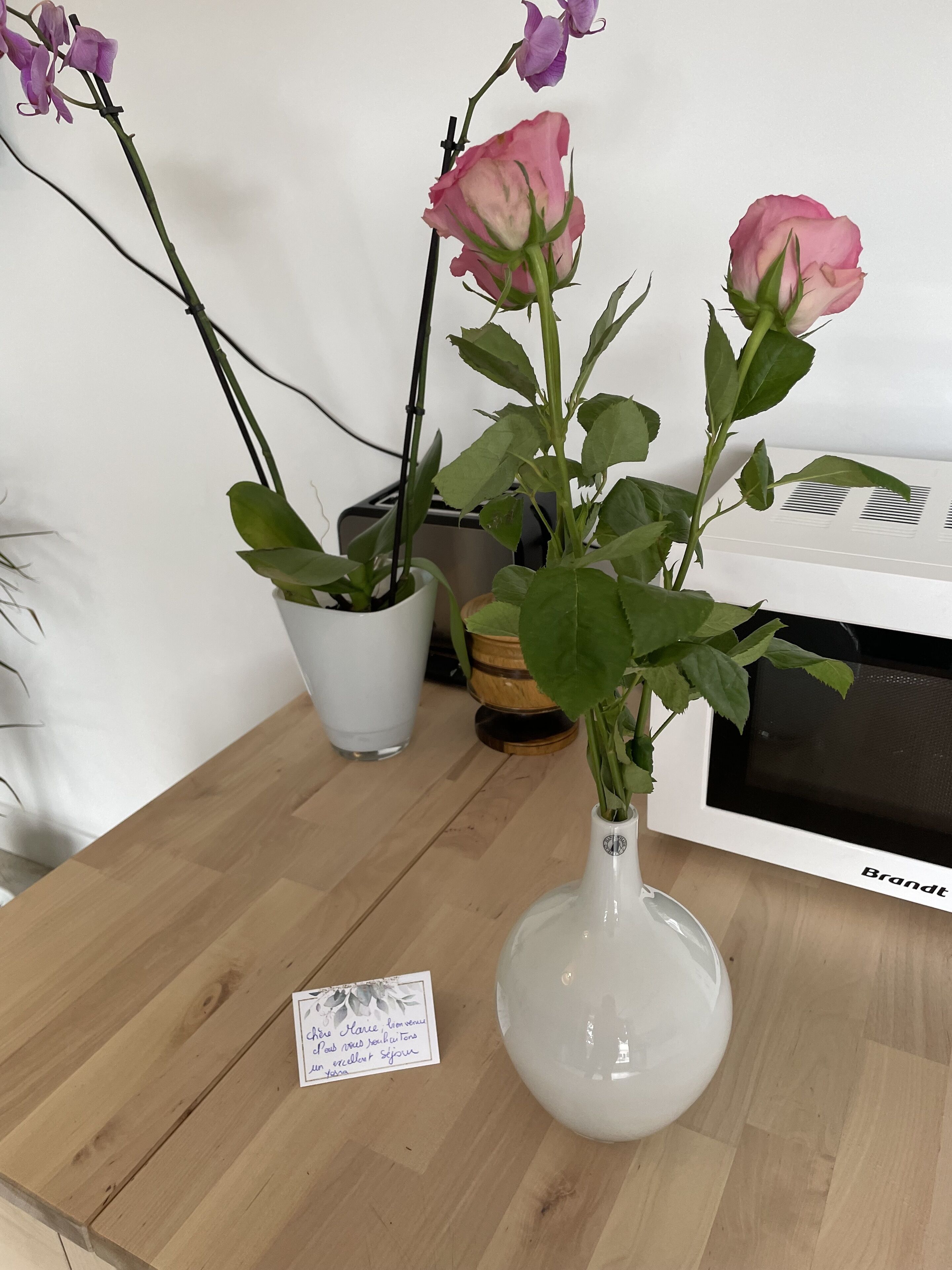 A wooden table with a white vase containing pink flowers, a white pot with green leaves, and a small card with text.