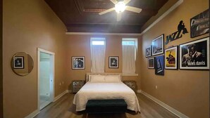 Un dormitorio con cama, ventilador de techo, cuadros enmarcados en las paredes y una puerta que da a otra habitación.