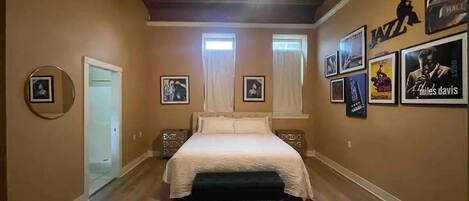 A bedroom with a bed, a ceiling fan, framed pictures on the walls, and a door leading to another room.