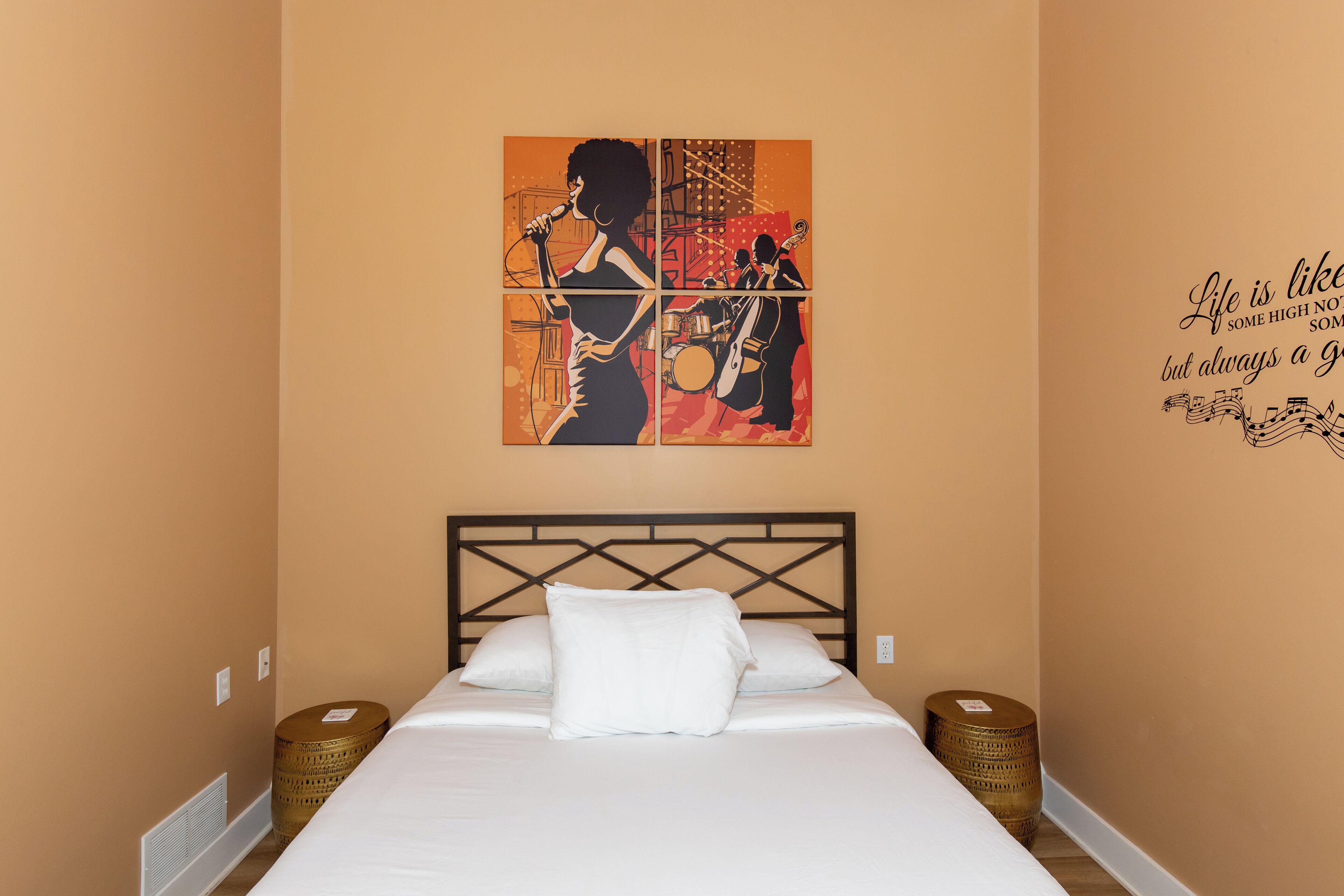 A neatly made bed with white linens and pillows, a headboard with a geometric pattern, and a wall with a musical-themed artwork and a motivational quote.
