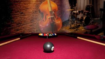 A pool table with a cue ball in the center, a pool rack in the background, and a brick wall with a mural of a violin.
