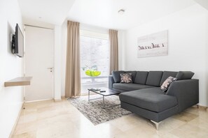 Un salon moderne avec un canapé d’angle gris, une table basse, un téléviseur à écran plat fixé au mur et une fenêtre avec des rideaux.