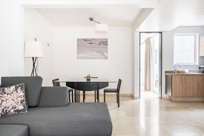 A modern living room with a grey sofa, a dining table with chairs, a lamp, and a kitchen area with cabinets and a window.