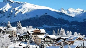 A snow-covered mountain resort with chalet-style buildings, a ski slope, and a forested backdrop.