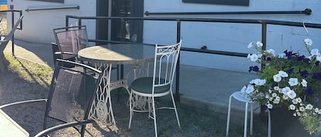 An outdoor seating area with a table and chairs, a small garden with flowers, and a building with a white exterior and black trim.