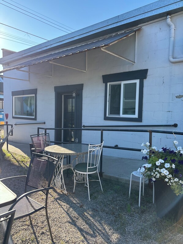An outdoor seating area with a table and chairs, a small garden with flowers, and a building with a white exterior and black trim. - Boutique Downtown Suite | Walk to Shops & Cafés | Sleeps 6 (Armstrong)