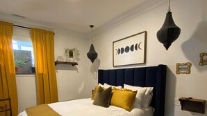 A neatly made bed with a white duvet and multiple pillows, a dark blue headboard, a black pendant light, and a framed artwork on the wall. - Boutique Downtown Suite | Walk to Shops & Cafés | Sleeps 6 (Armstrong)