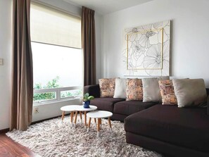 Un salon avec un canapé de couleur foncée, des coussins à motifs, une petite table blanche ornée d’une plante et une grande fenêtre avec des rideaux.
